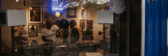 Zwei Personen stehen in einem Tonstudio in einem Container. Das Bild wird durch eine Scheibe gemaQcht. Im Hintergrund an der Wand ist das Jägermeister Logo und der Schriftzug "Studio CanV"